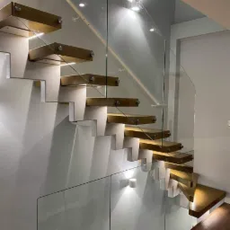 Nowoczesne schody wspornikowe z drewnianymi stopniami, mocowane do ściany, z przeszkloną balustradą, oświetlone punktowymi lampami w ścianie.