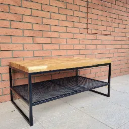 Stolik kawowy z blatem z jasnego drewna i metalową konstrukcją z siatkową półką, ustawiony na jasnych płytkach na tle ściany z cegły klinkierowej z widocznymi elementami instalacji.