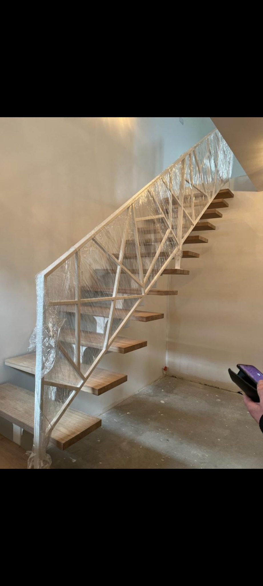 Nowoczesne schody z drewnianymi stopniami i metalową balustradą o geometrycznym wzorze, zabezpieczoną folią ochronną, w surowym wnętrzu.