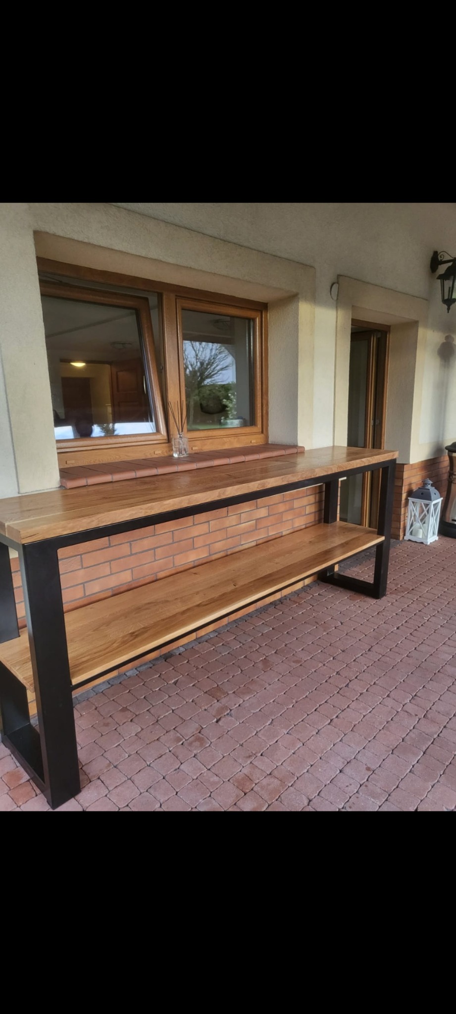 Drewniana półka z czarną metalową ramą na zewnątrz budynku w Zabierzowie, widoczna ceglana ściana w tle i kostka brukowa na podłodze.