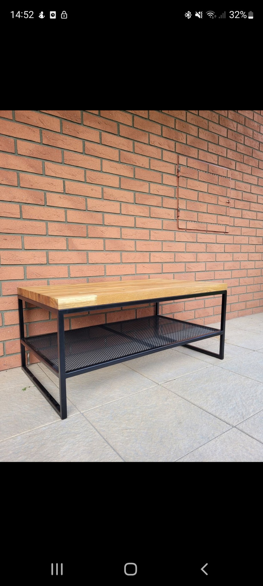 Stolik kawowy z blatem z jasnego drewna i metalową konstrukcją z siatkową półką, ustawiony na jasnych płytkach na tle ściany z cegły klinkierowej z widocznymi elementami instalacji.