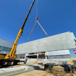 Żółty dźwig podnosi betonowy panel ścienny na placu budowy, widoczny fragment powstającego budynku z otworami okiennymi, stos paneli na ziemi, znak zakazu w tle.
