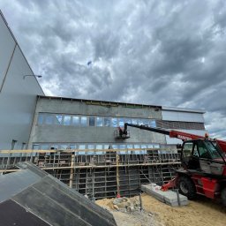 Axis Projekt Paweł Kozłowski - Budowa budynku przemysłowego: pracownik w podnośniku koszowym na tle szarej elewacji z oknami, widoczne szalunki i czerwony dźwig Manitou, pochmurne niebo