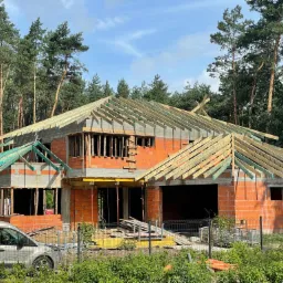 Surowy stan budynku z czerwonej cegły otoczonego lasem, widoczne drewniane elementy konstrukcji dachu i szalunki okienne, srebrny van parkuje przed ogrodzeniem.