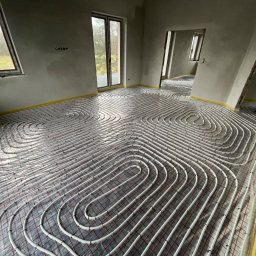 Axis Projekt Paweł Kozłowski - Rozłożona instalacja ogrzewania podłogowego w surowym wnętrzu z widocznymi oknami i drzwiami balkonowymi, zabezpieczona folią i taśmą na obrzeżach pomieszczenia.