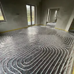 Rozłożona instalacja ogrzewania podłogowego w surowym wnętrzu z widocznymi oknami i drzwiami balkonowymi, zabezpieczona folią i taśmą na obrzeżach pomieszczenia.