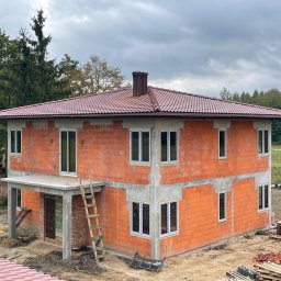 Axis Projekt Paweł Kozłowski - Dwukondygnacyjny dom w trakcie budowy, ściany z czerwonej cegły, zamontowane okna PCV, brązowy dach z kominem, drewniana drabina oparta o ścianę, teren budowy z paletami i gruzem, w tle drzewa...