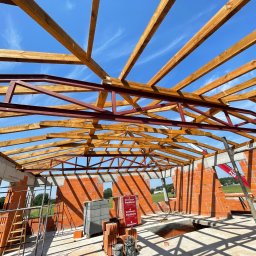 Axis Projekt Paweł Kozłowski - Wnętrze domu w trakcie budowy: murowane ściany z czerwonej cegły Porotherm, drewniana konstrukcja dachu z elementami stalowymi, błękitne niebo widoczne przez otwory okienne, paleta cegieł i drabina...