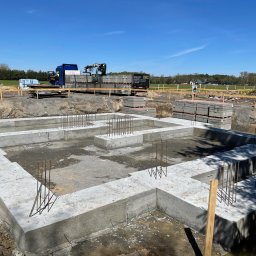 Axis Projekt Paweł Kozłowski - Wylane betonowe fundamenty domu jednorodzinnego z wystającymi prętami zbrojeniowymi, na działce budowlanej, w tle ciężarówka z dźwigiem i palety z bloczkami betonowymi.