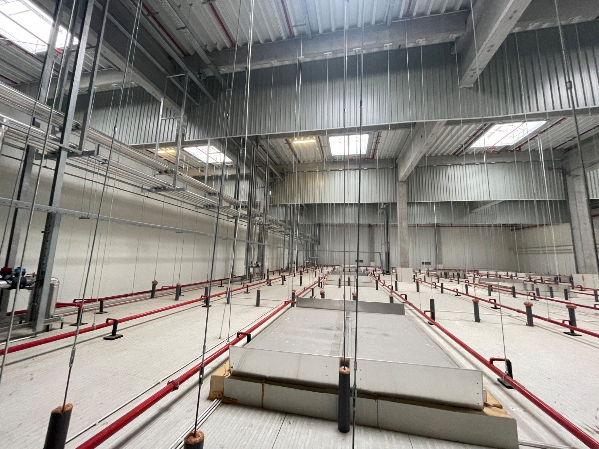 Wnętrze hali przemysłowej w trakcie budowy, widoczne instalacje pod sufitem, rury w kolorze czerwonym i metalowe elementy konstrukcyjne, oświetlenie naturalne przez okna w dachu.