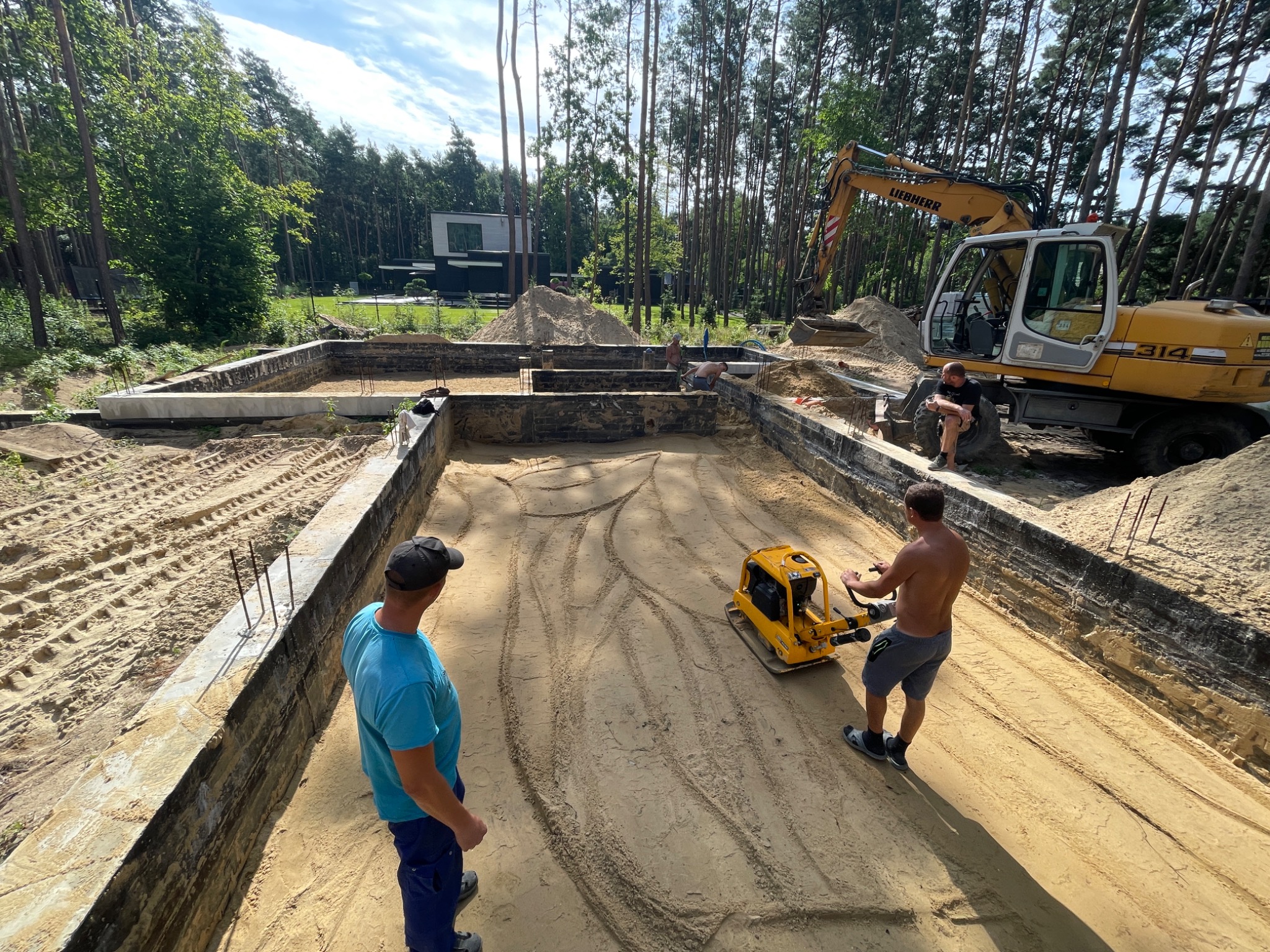 Wyrównywanie piasku wewnątrz fundamentów pod budowę domu jednorodzinnego z użyciem zagęszczarki wibracyjnej, w tle koparka Liebherr i budynek w otoczeniu drzew.