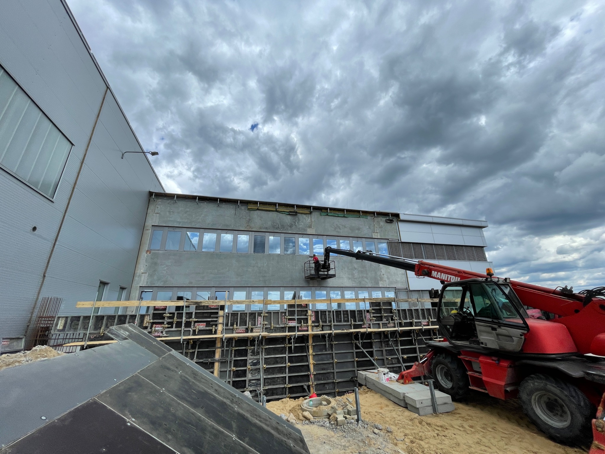 Budowa budynku przemysłowego: pracownik w podnośniku koszowym na tle szarej elewacji z oknami, widoczne szalunki i czerwony dźwig Manitou, pochmurne niebo