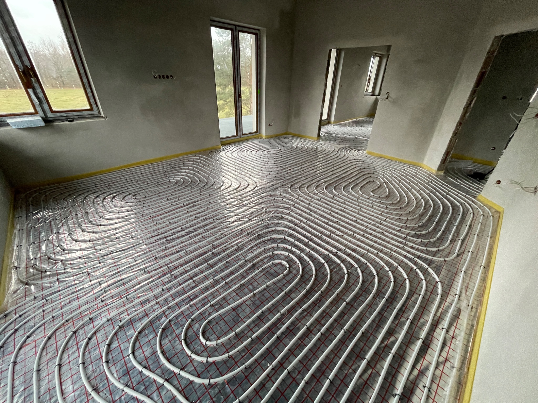 Rozłożona instalacja ogrzewania podłogowego w surowym wnętrzu z widocznymi oknami i drzwiami balkonowymi, zabezpieczona folią i taśmą na obrzeżach pomieszczenia.