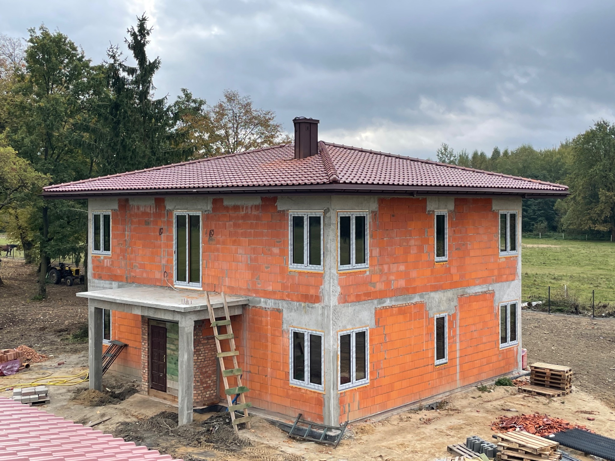 Dwukondygnacyjny dom w trakcie budowy, ściany z czerwonej cegły, zamontowane okna PCV, brązowy dach z kominem, drewniana drabina oparta o ścianę, teren budowy z paletami i gruzem, w tle drzewa...