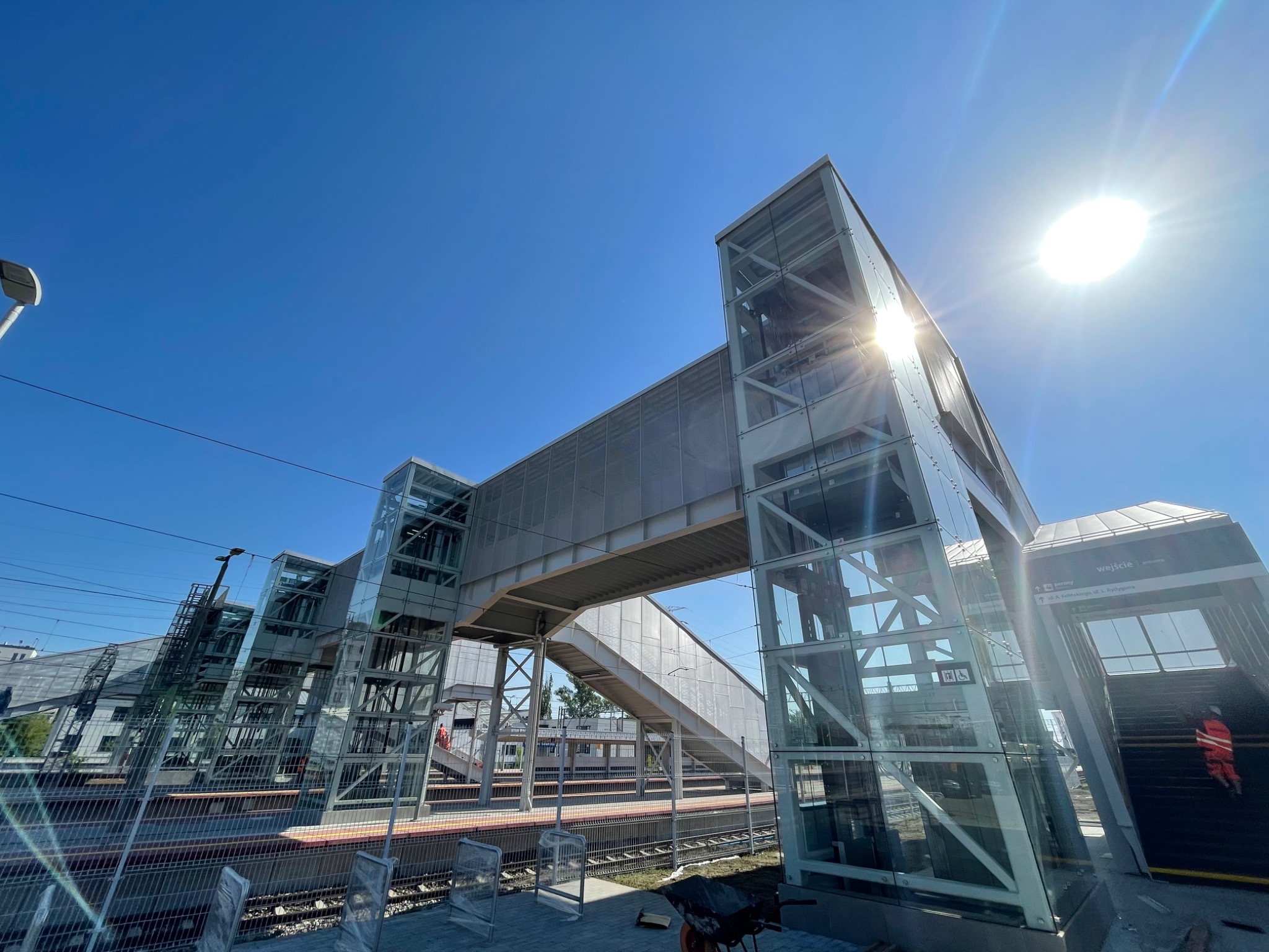 Nowoczesna kładka dla pieszych nad peronami kolejowymi, wykonana ze stali i szkła, w słoneczny dzień.