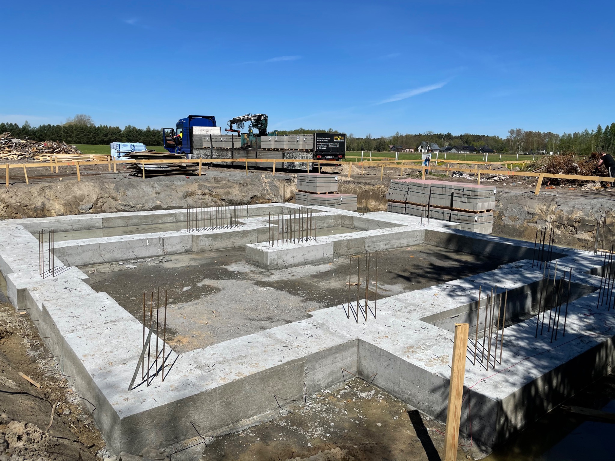 Wylane betonowe fundamenty domu jednorodzinnego z wystającymi prętami zbrojeniowymi, na działce budowlanej, w tle ciężarówka z dźwigiem i palety z bloczkami betonowymi.