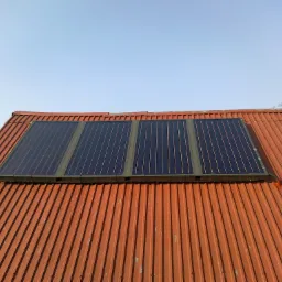 Cztery panele słoneczne zamontowane na dachu z blachy falistej w kolorze ceglastym, widoczne z perspektywy dachu pod lekkim kątem, na tle błękitnego nieba.