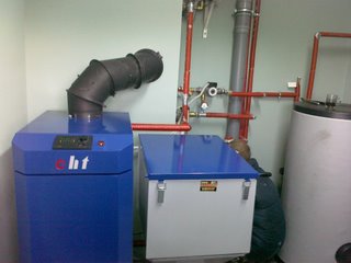 Instalacja kotła grzewczego w kotłowni, widoczne rury miedziane, komin i zbiornik na wodę. Pracownik w trakcie montażu, częściowo zasłonięty przez urządzenia.