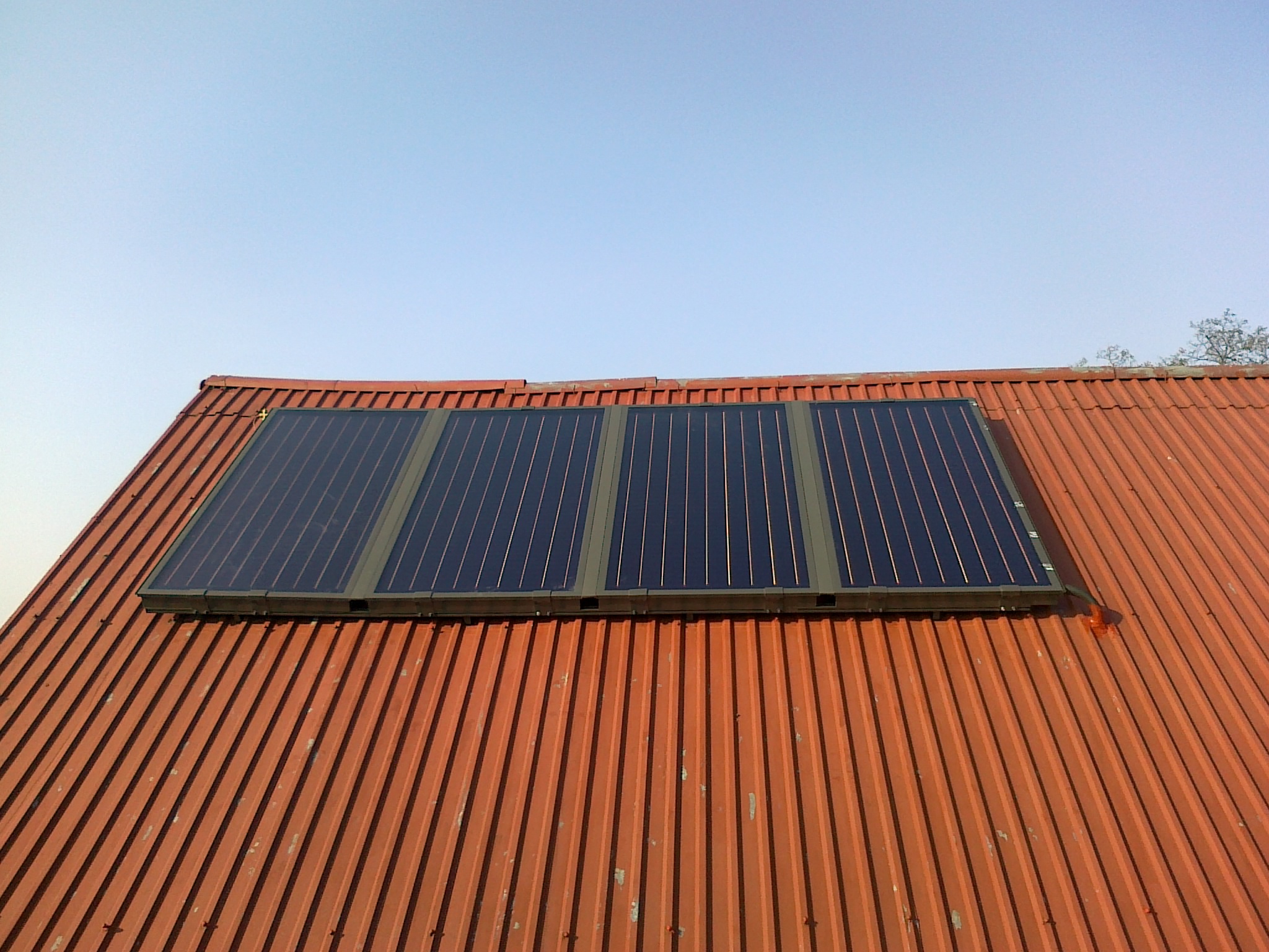 Cztery panele słoneczne zamontowane na dachu z blachy falistej w kolorze ceglastym, widoczne z perspektywy dachu pod lekkim kątem, na tle błękitnego nieba.