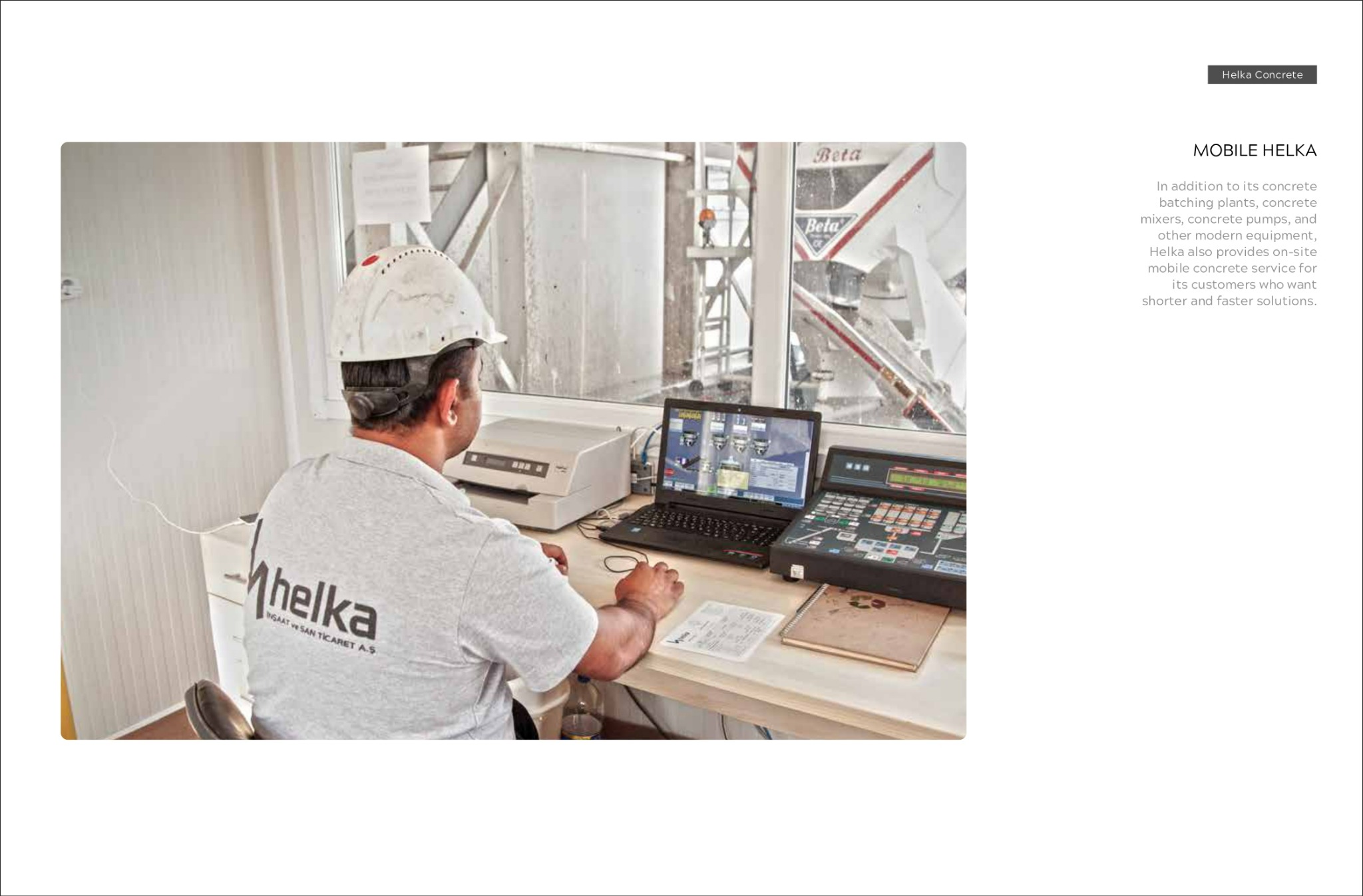 Pracownik w białym kasku obsługuje komputer sterujący betonownią, widoczna maszyna betonowa za oknem i logo firmy na koszulce