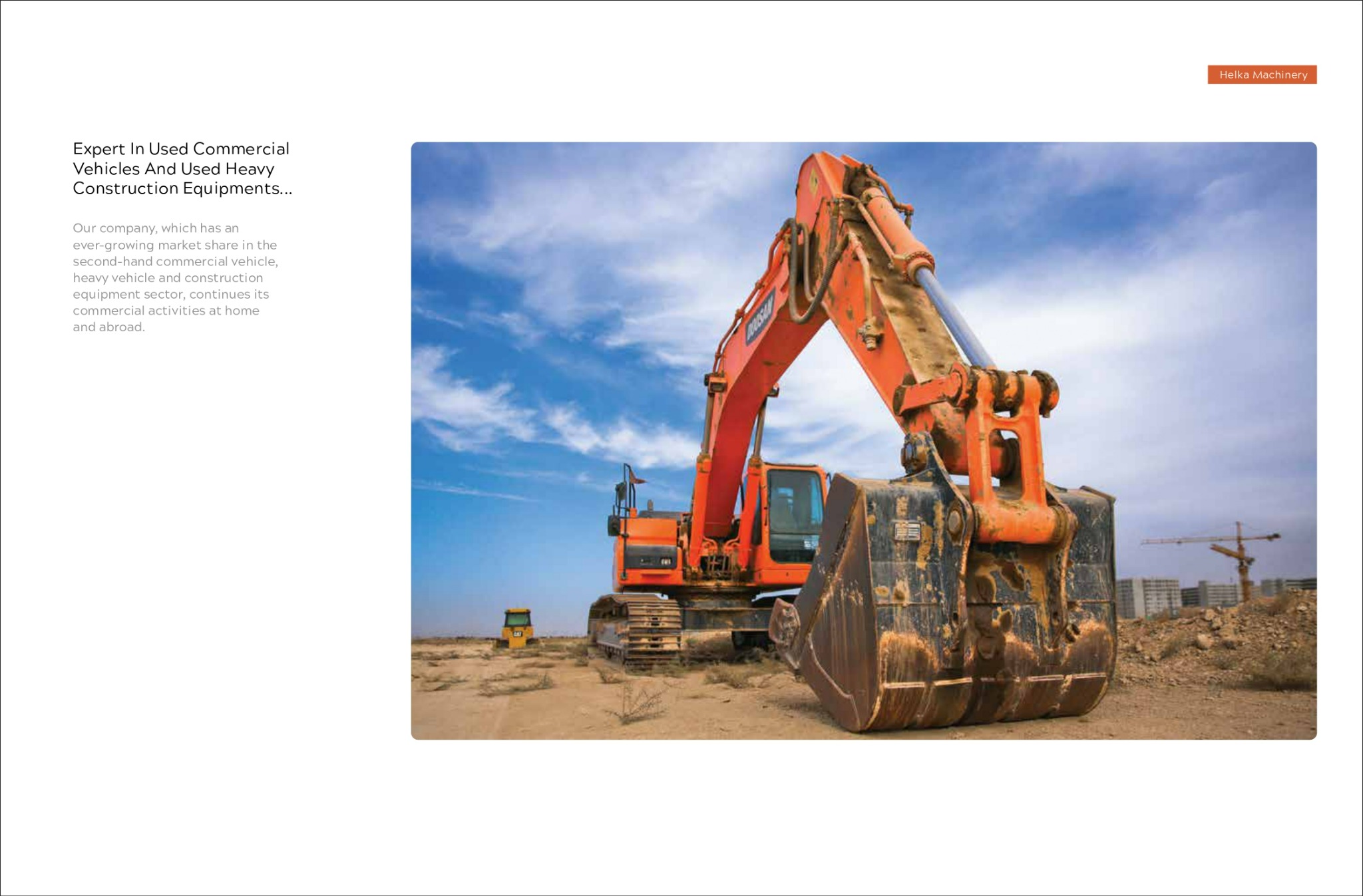 Pomarańczowa koparka na placu budowy, z widocznymi śladami użytkowania na łyżce, w tle zabudowa miejska i żuraw budowlany pod błękitnym niebem.