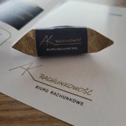 Cukierek z logo 'AK Rachunkowość Biuro Rachunkowe' leżący na papierze firmowym z tym samym logo, otwarty folder w tle.