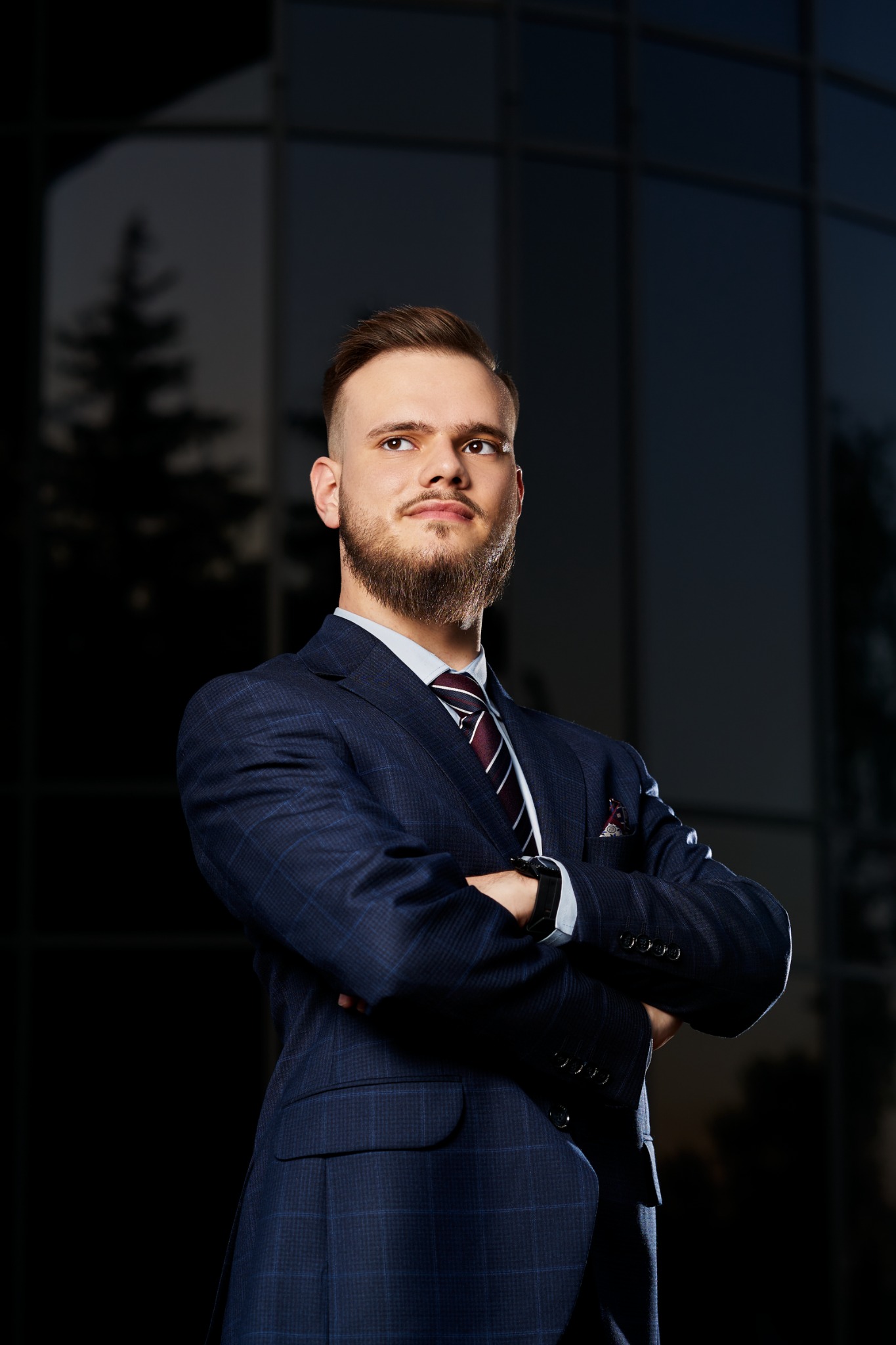 Portret brodatego mężczyzny w niebieskim garniturze w kratę, z założonymi rękami na tle nowoczesnej szklanej fasady budynku.