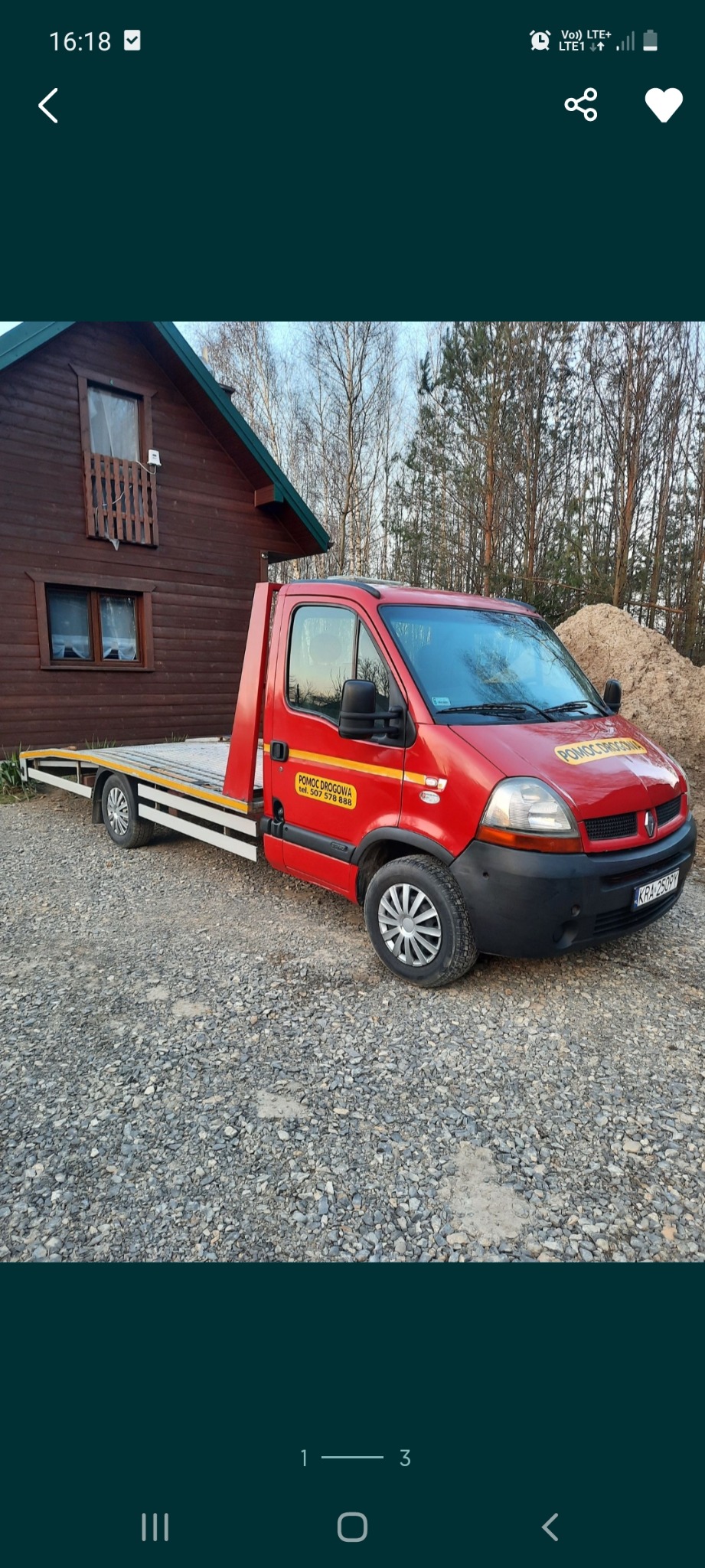 Czerwona laweta Renault Master z żółtymi napisami 'POMOC DROGOWA' zaparkowana na żwirowym podłożu przed drewnianym domkiem z poddaszem i stertą piasku w tle.