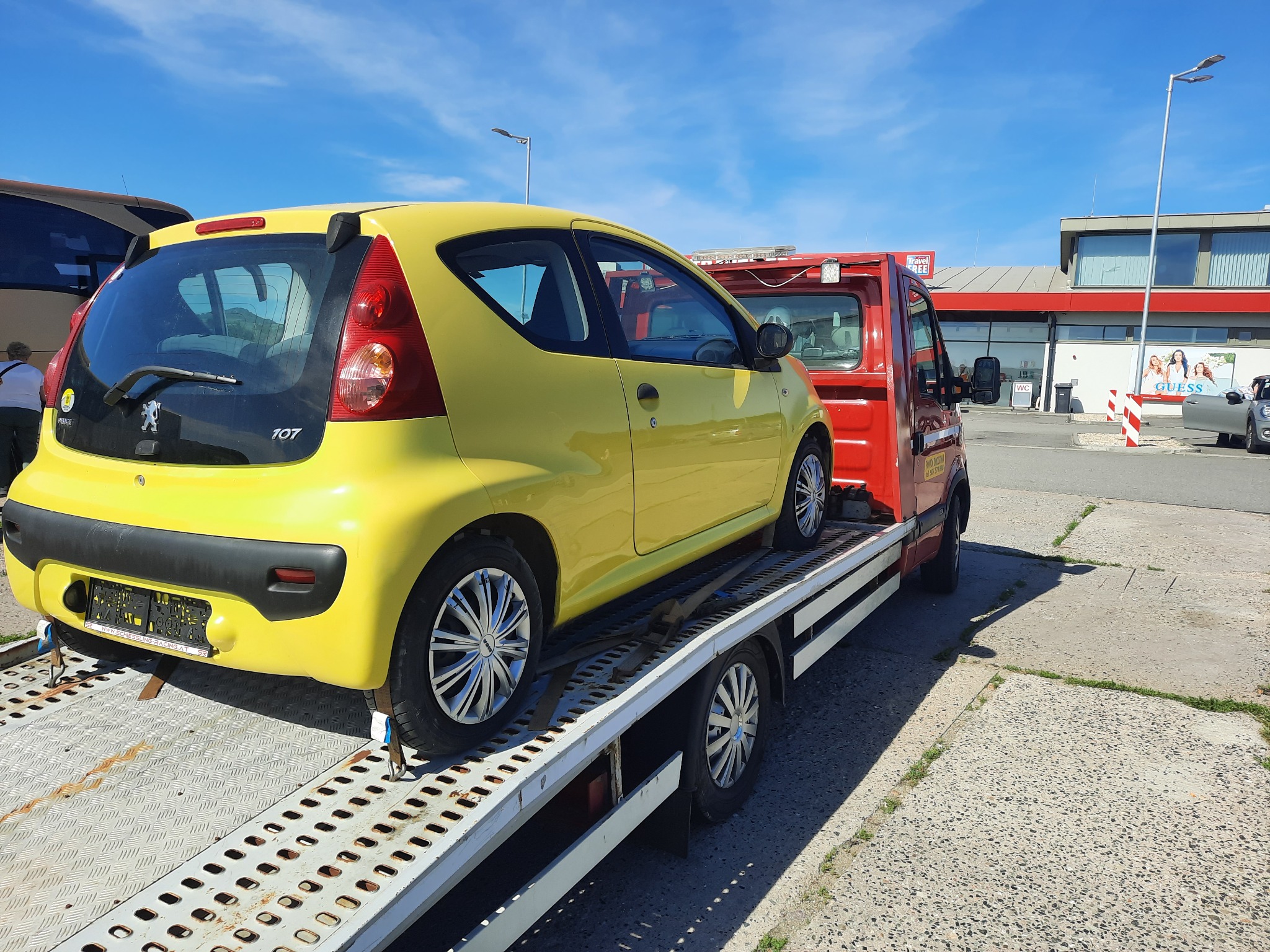 Żółty samochód osobowy Peugeot 107 zabezpieczony pasami na platformie czerwonej lawety, na parkingu przed budynkiem z szyldem Guess.