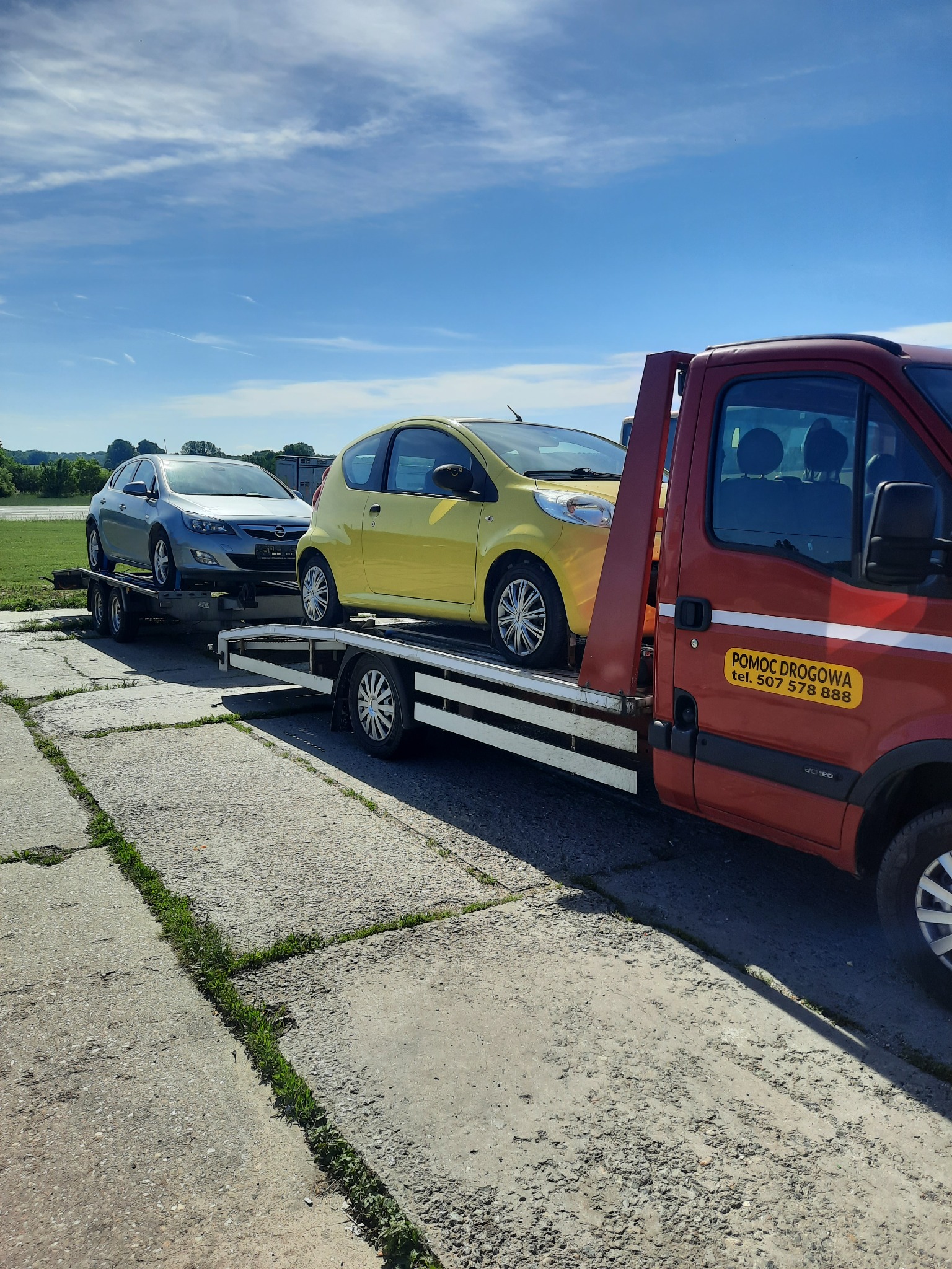 Czerwona laweta z żółtym samochodem osobowym i srebrnym kombi na platformie, widoczny napis 'Pomoc Drogowa' na drzwiach kabiny, betonowa nawierzchnia z trawą w tle.