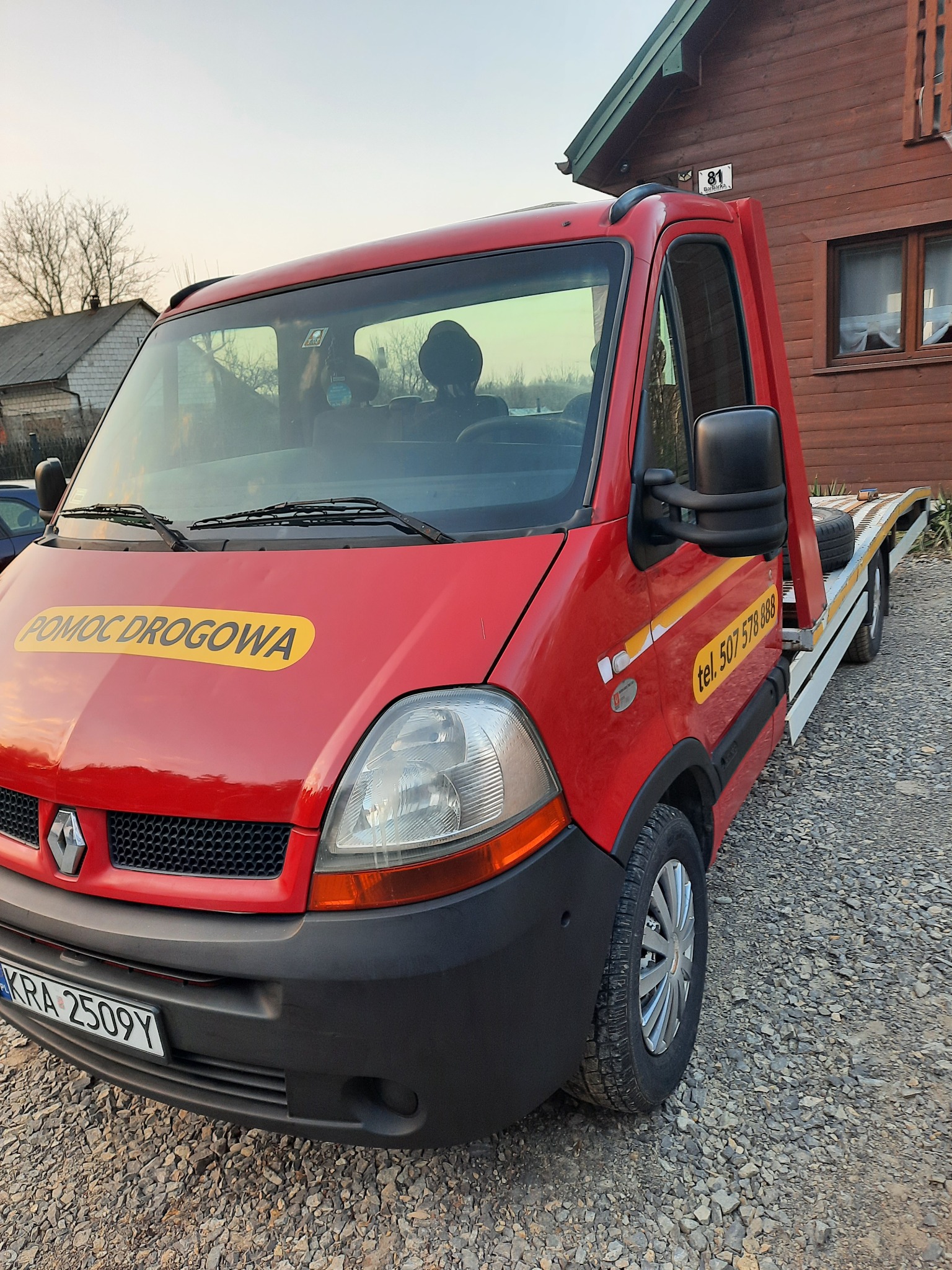 Czerwona laweta Renault Master z żółtym napisem 'POMOC DROGOWA' i numerem telefonu, zaparkowana na żwirowym podłożu przed drewnianym domem z numerem 81.