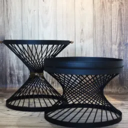 Dwa czarne stoliki kawowe o geometrycznej konstrukcji z metalowych prętów, ustawione na drewnianej podłodze na tle drewnianej ściany.