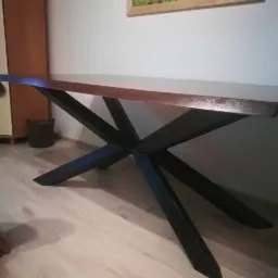 Stół z blatem z ciemnego drewna i geometryczną, czarną podstawą z metalowych profili na jasnej podłodze, w tle ściana z obrazem górskiego krajobrazu.