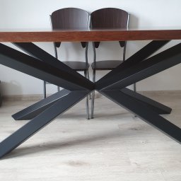 Metal&Wood Art Maciej Gierszewski - Stół z blatem z ciemnego drewna i czarną, metalową podstawą w kształcie litery X, widziane z dołu, z dwoma krzesłami w tle i koszem z lewej strony.