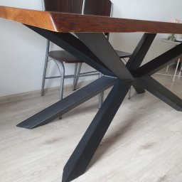 Metal&Wood Art Maciej Gierszewski - Stół z blatem z jasnego drewna i czarną, metalową podstawą w kształcie gwiazdy, widoczne krzesła w tle.