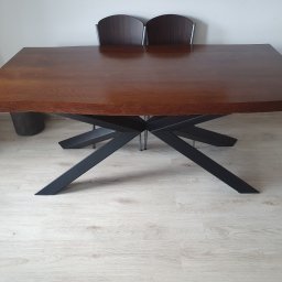 Metal&Wood Art Maciej Gierszewski - Stół z blatem z ciemnego drewna o nieregularnych krawędziach, wsparty na czarnej, geometrycznej podstawie, ustawiony na jasnej podłodze, z dwoma krzesłami w tle.