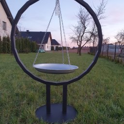 Metal&Wood Art Maciej Gierszewski - Metalowa konstrukcja w kształcie okręgu z podwieszaną misą na łańcuchach, stojąca na trawniku przed domem jednorodzinnym o zmierzchu.