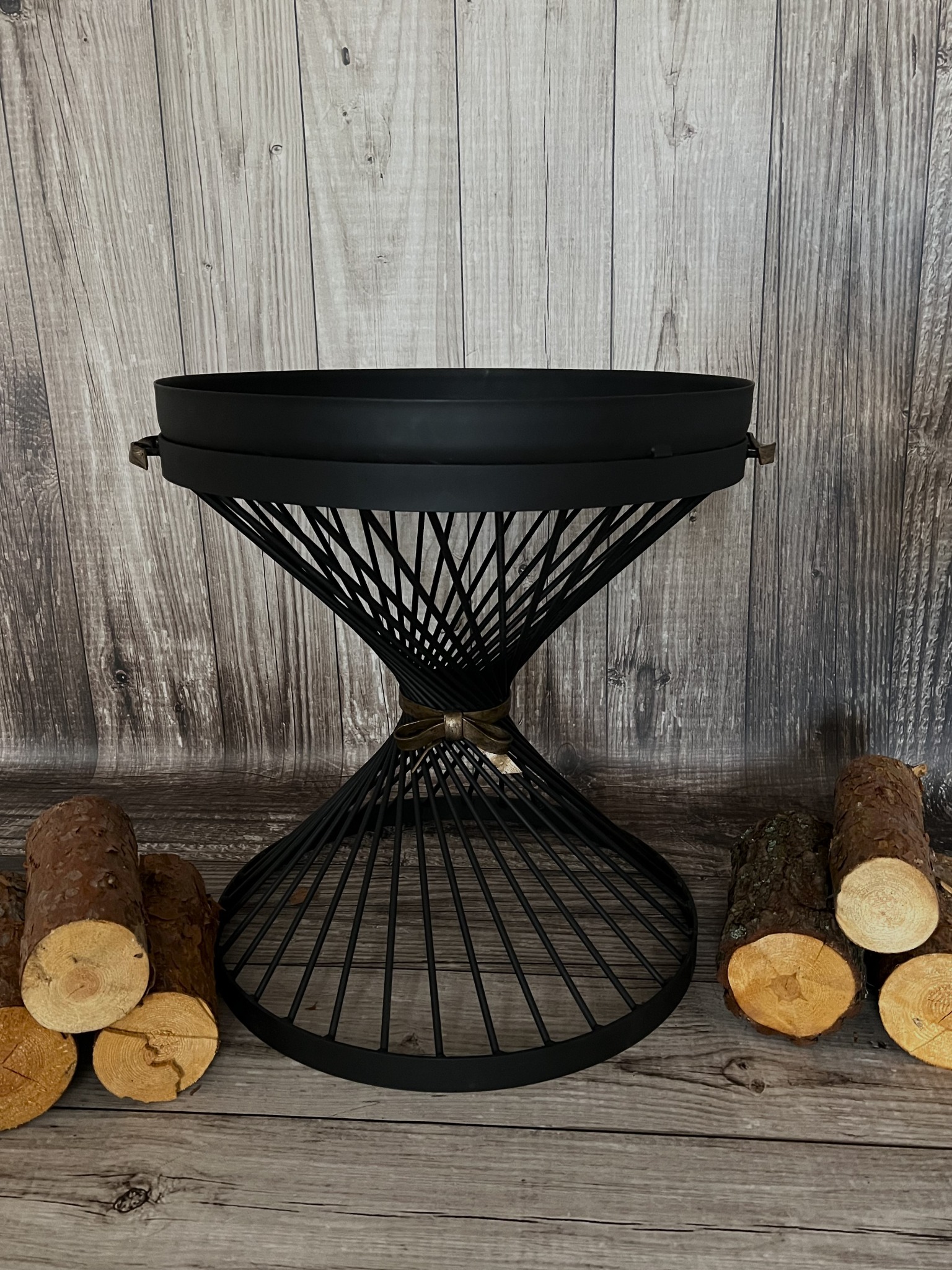 Czarny, metalowy kwietnik o geometrycznej konstrukcji z drewnianym tłem i dekoracyjnymi kawałkami drewna obok.