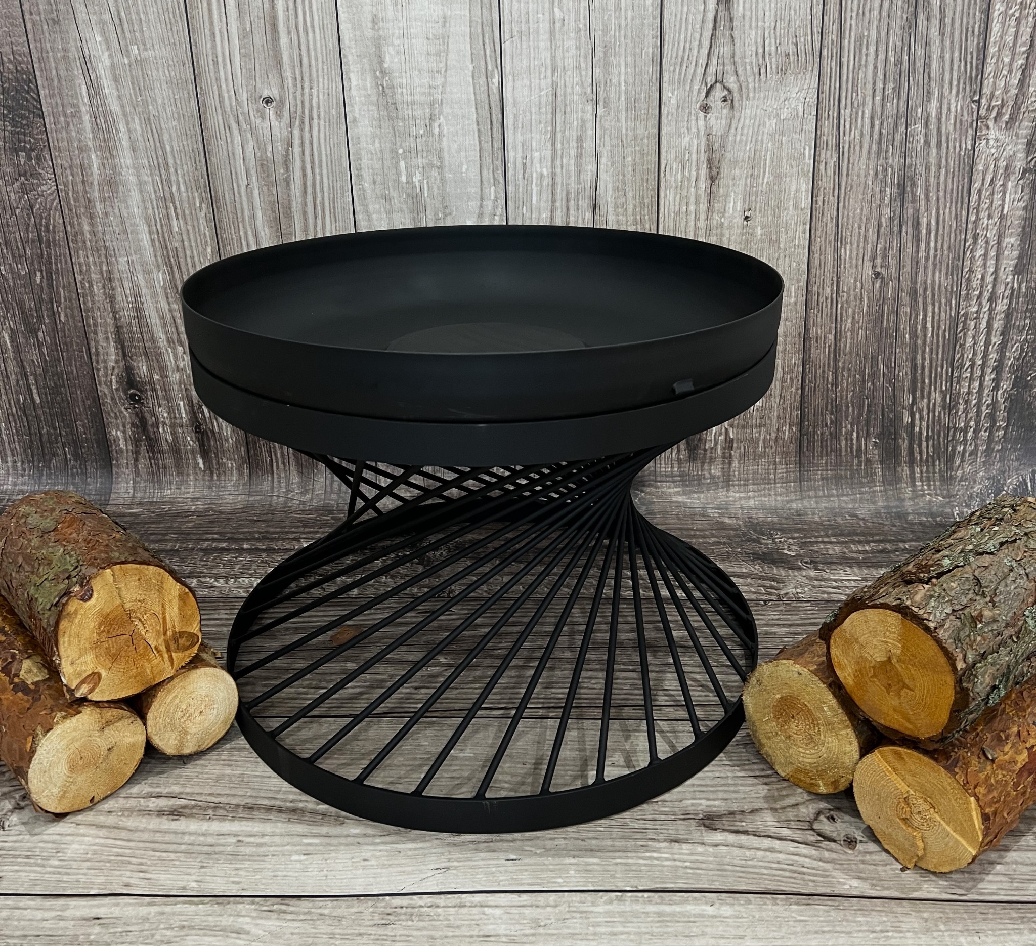 Czarny, metalowy kosz na ognisko z spiralną podstawą, otoczony ułożonymi kawałkami drewna na tle drewnianej ściany.