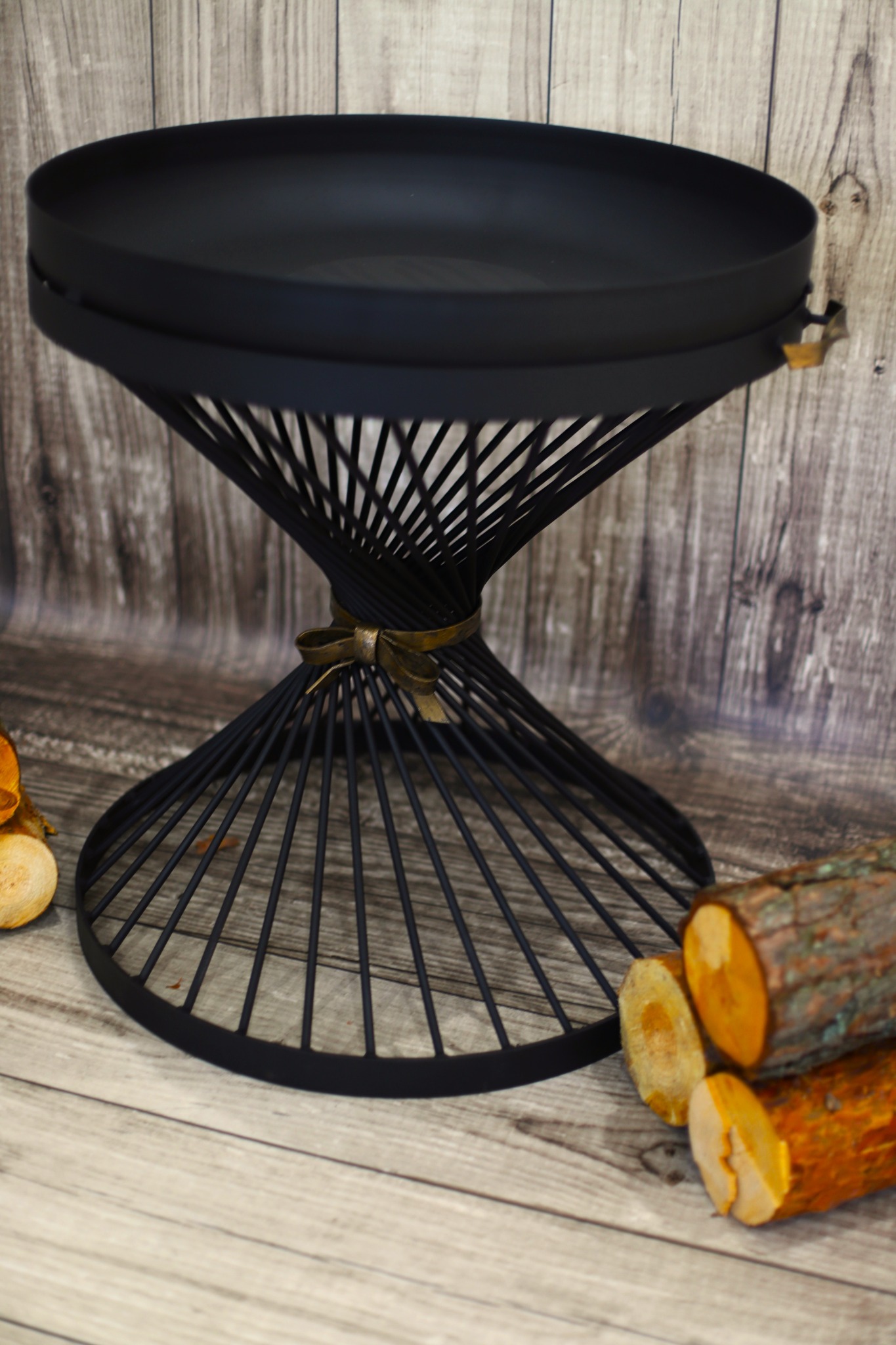 Czarny, metalowy stolik ogrodowy z dekoracyjną podstawą w kształcie klepsydry, ozdobiony złotą wstążką, obok ułożone kawałki drewna na tle drewnianej ściany.