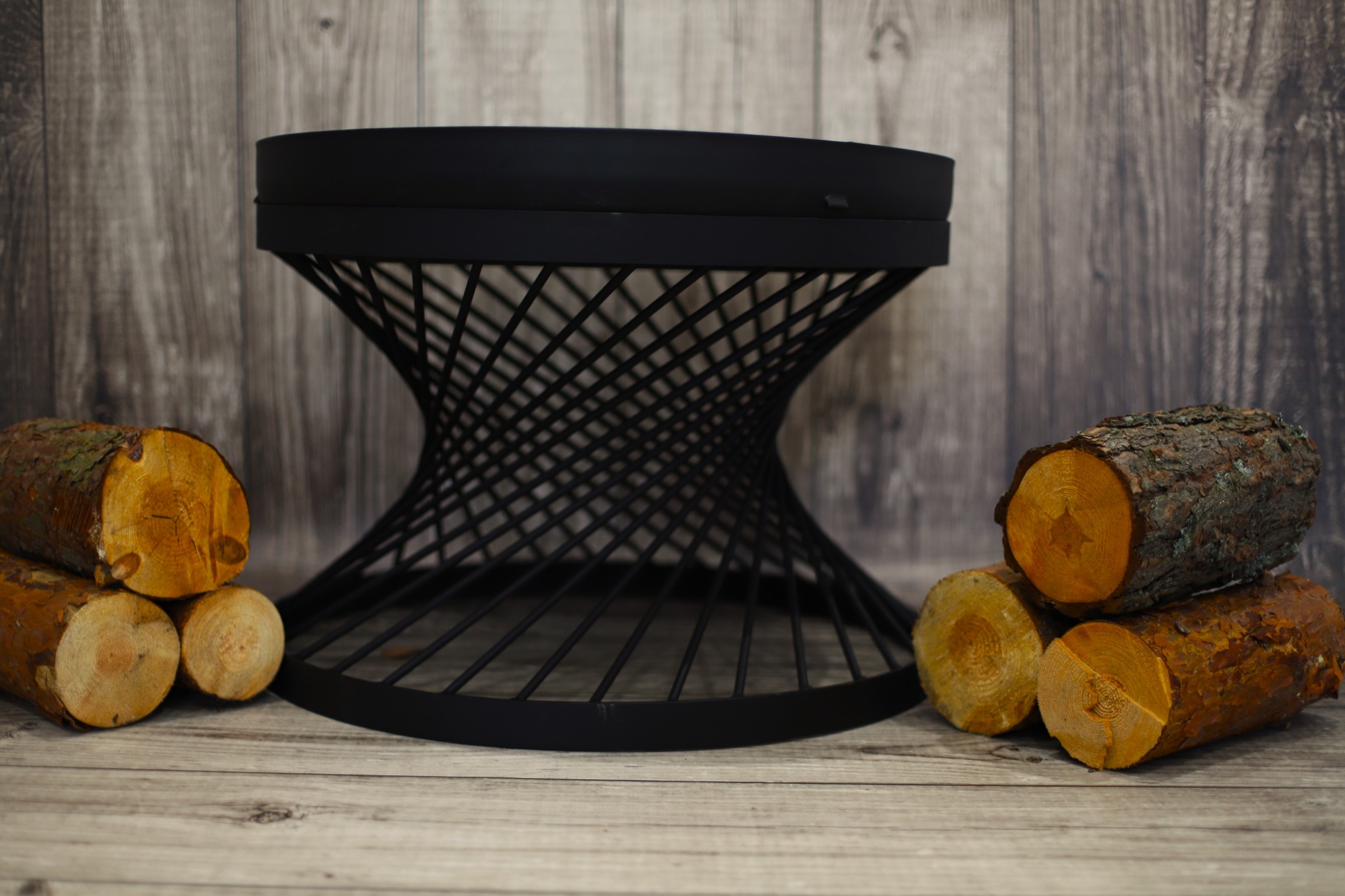 Metalowy kosz na drewno kominkowe o spiralnej konstrukcji, otoczony ułożonymi kłodami drewna na drewnianym tle.