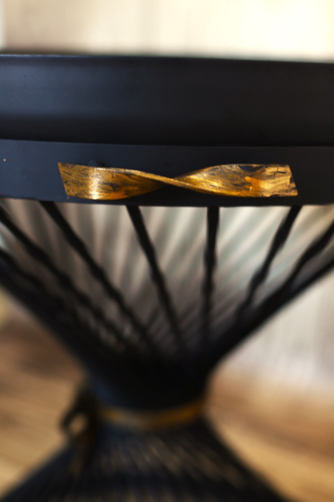 Fragment czarnej metalowej konstrukcji meblowej z dekoracyjnym, złotym elementem w kształcie skręconej wstążki, widoczne pionowe pręty w tle.