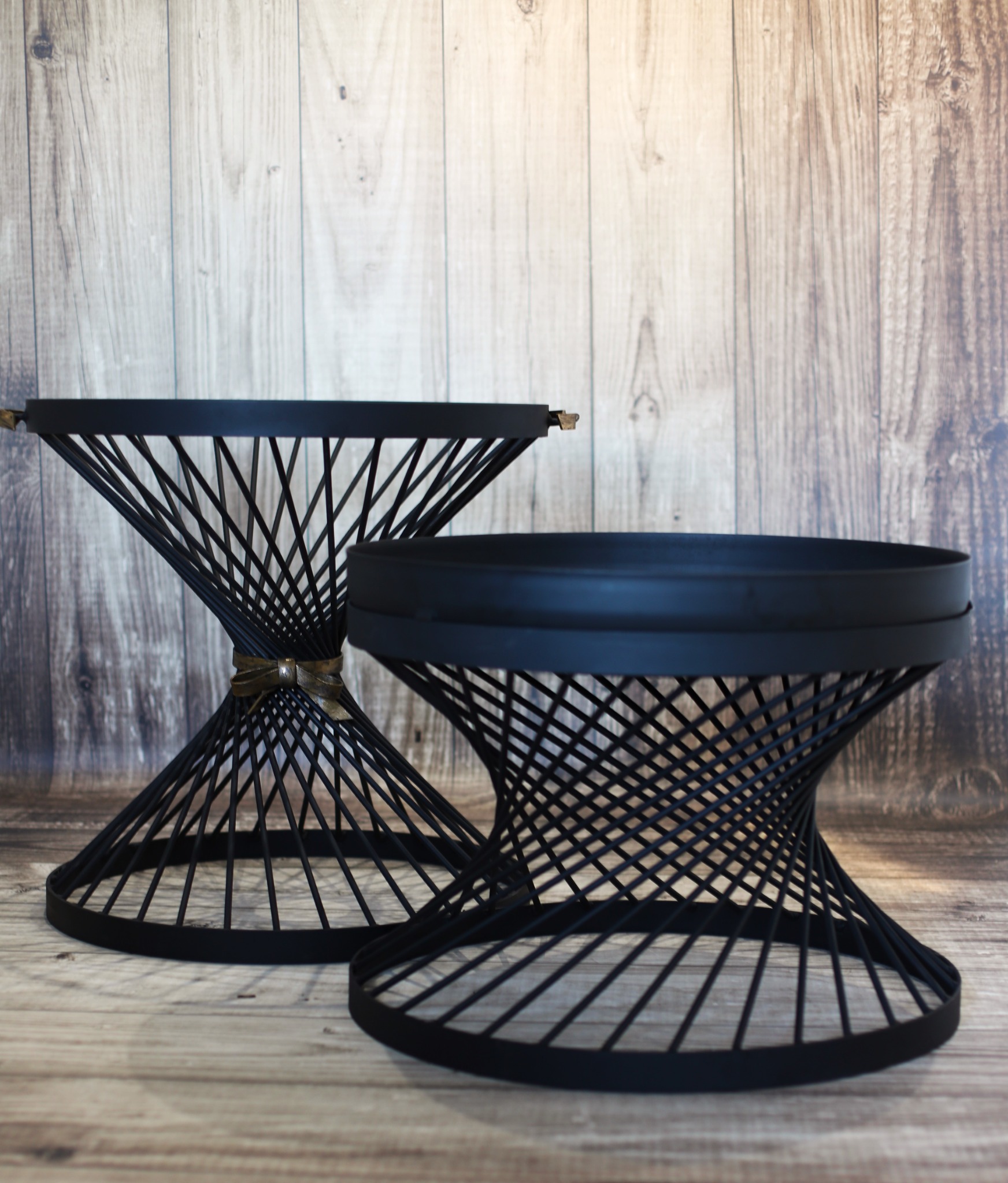 Dwa czarne stoliki kawowe o geometrycznej konstrukcji z metalowych prętów, ustawione na drewnianej podłodze na tle drewnianej ściany.