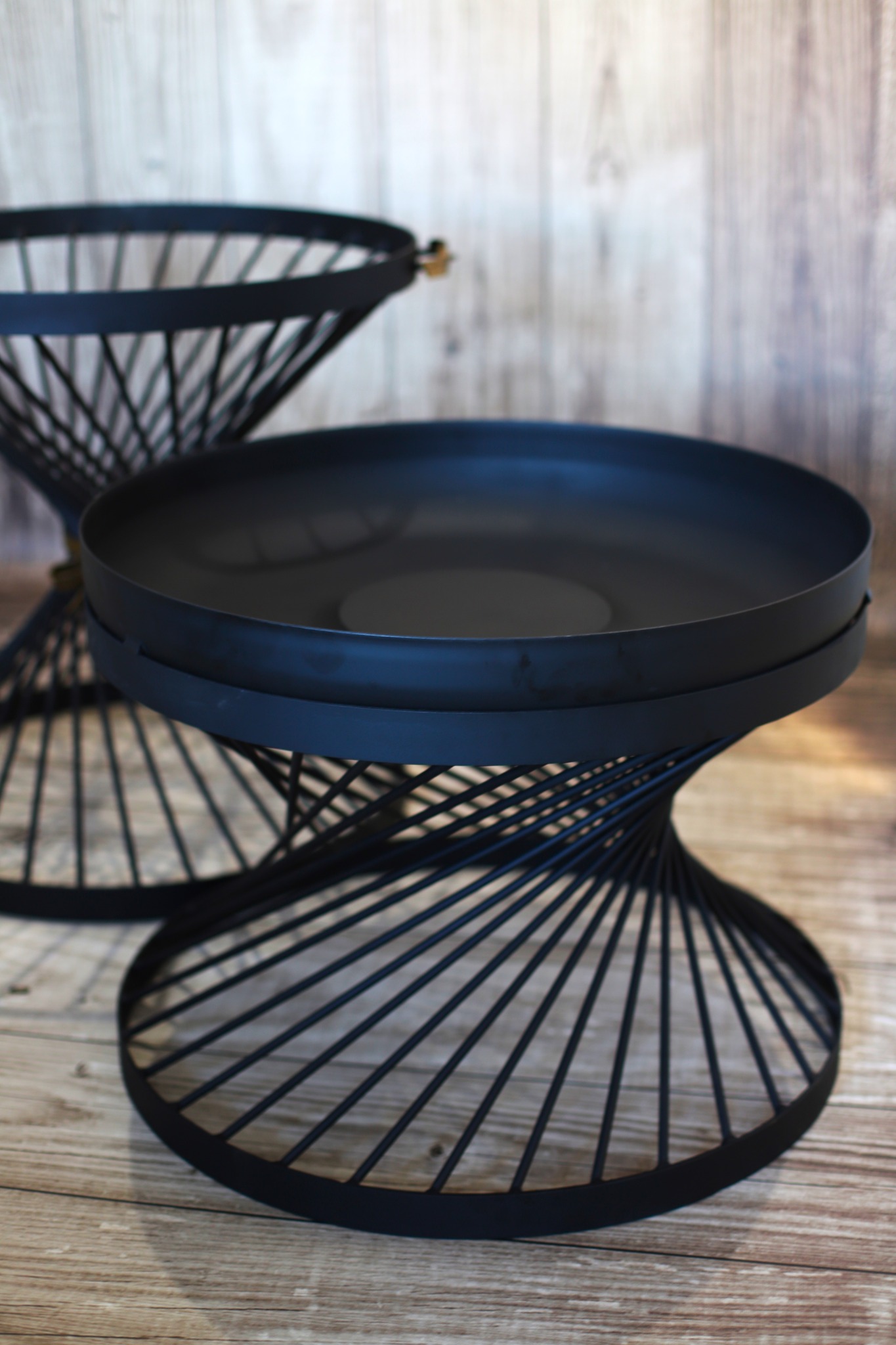 Dwie metalowe, czarne podstawki z okrągłymi blatami i spiralnymi nogami ustawione na drewnianej podłodze na tle drewnianej ściany.