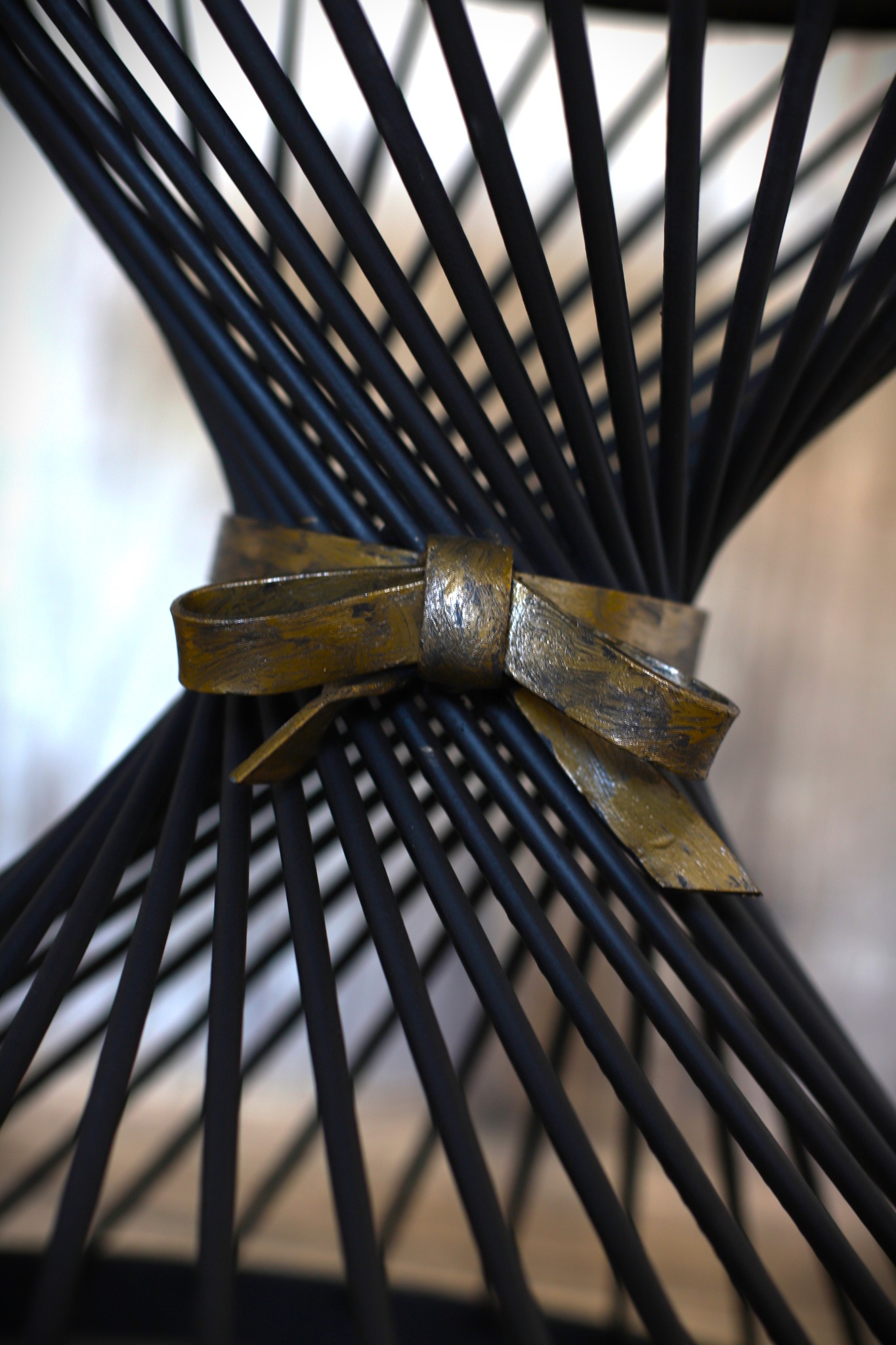 Abstrakcyjna konstrukcja z czarnych prętów spiętych metalową kokardą z efektem postarzenia, ukazana z bliska w miękkim oświetleniu.