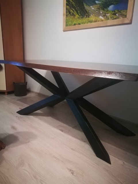 Stół z blatem z ciemnego drewna i geometryczną, czarną podstawą z metalowych profili na jasnej podłodze, w tle ściana z obrazem górskiego krajobrazu.