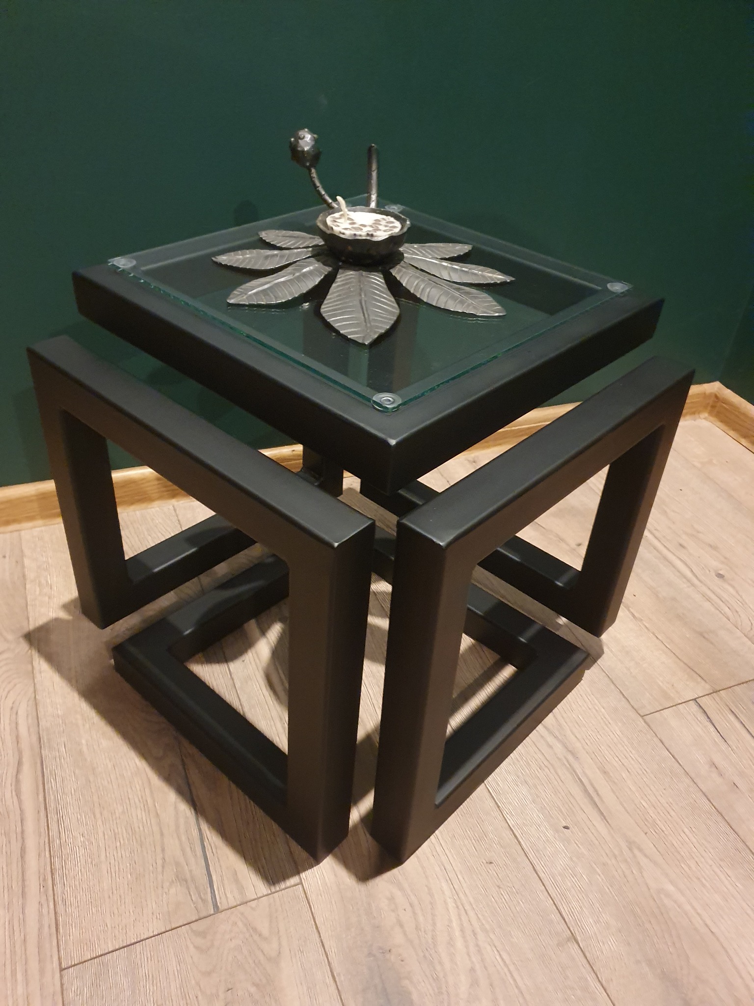 Czarny, metalowy stolik kawowy o geometrycznej konstrukcji z blatem ze szkła, ozdobiony metalową kompozycją przypominającą kwiat.