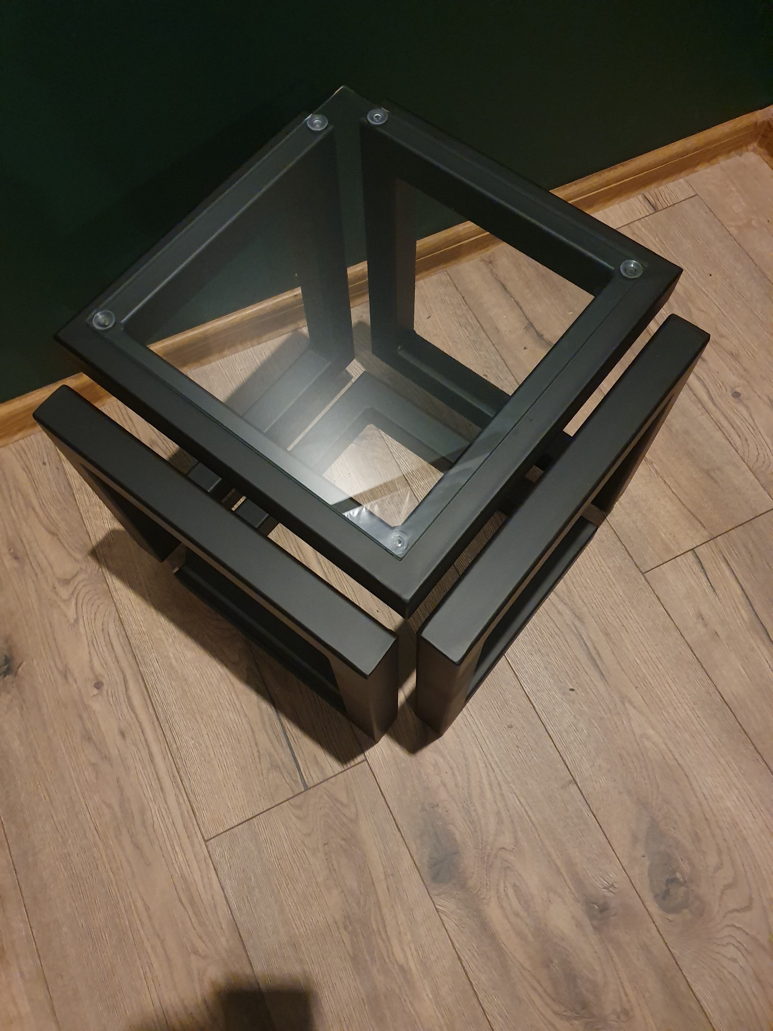 Kwadratowy stolik kawowy z czarną, metalową ramą i szklanym blatem, widoczny na tle drewnianej podłogi i ciemnozielonej ściany.
