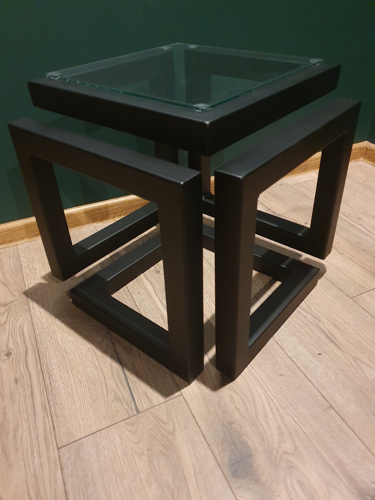 Nowoczesny stolik kawowy z czarną, metalową podstawą w geometrycznym stylu i szklanym blatem na tle ciemnozielonej ściany i drewnianej podłogi.
