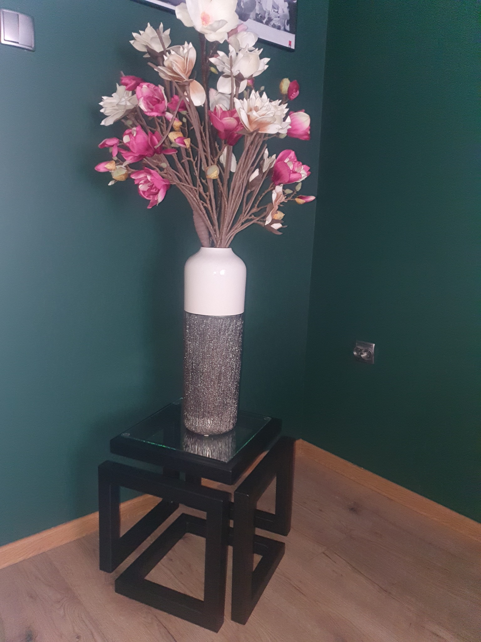 Dekoracyjny bukiet z różowymi i białymi kwiatami w wazonie na nowoczesnym stoliku z czarnymi, geometrycznymi nogami, na tle ciemnozielonej ściany.